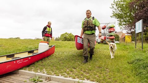 Zwei Personen und ein Hund am Flussufer mit einem Kanu