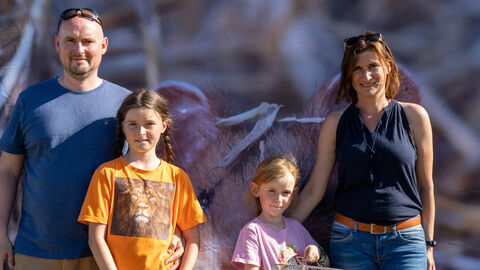 Eine Familie steht vor einem Feldhamsterplakat. Das eine Mädchen hält einen Käfig mit einem Feldhamster