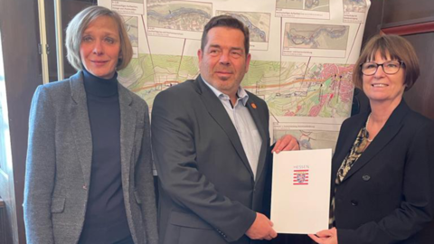 Drei Personen stehen vor einer Wand. Ministerin Hinz übergibt einen Bescheid