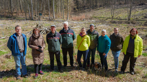 9 Personen stehen im Wald. Im Hintergrund Teil Gehölz, die Bäume haben noch keine Blätter.