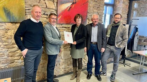 Vier Männer und eine Frau stehen vor einer Steinwand. Die Frau übergibt eine Urkunde.