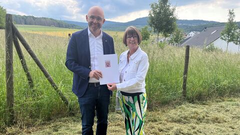 Bildtext: Kommunales Bodenschutzkonzept. 75.000 Euro Förderung für Idstein. Zwei Personen (links Bürgermeister Christian Herfurth, rechts Ministerin Priska Hinz) stehen in der Natur. Gemeinsam halten sie einen weißen Umschlag mit Hessen Wappen in den Händen.