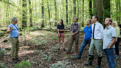 Feierliche Begehung des Naturschutzgebiets Gruppenbild im Wald. Eine Person erklärt die anderen hören zu.