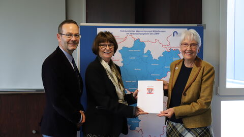 Ein Mann und zwei Frauen stehen vor einer Stellwand. Die Frauen halten einen Förderbescheid in der Hand