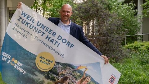 Minister Jung präsentiert neuen Banner für den Dorfwettbewerb 2024/25 Minister Jung hält Banner in der Hand. Darauf steht "Weil unser Dorf Zukunft hat! Hessischer Dorfwettbewerb 2024/2025