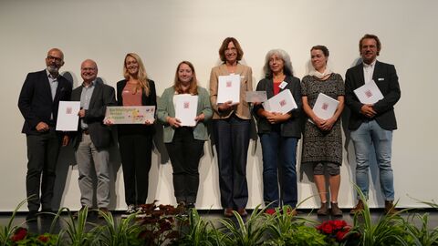 Staatssekretär Daniel Köfer (l.) zusammen mit Vertreterinnen und Vertretern der Umweltzentren, die die Urkunden stellvertretend für die Schulen auf der Bühne entgegennahmen.